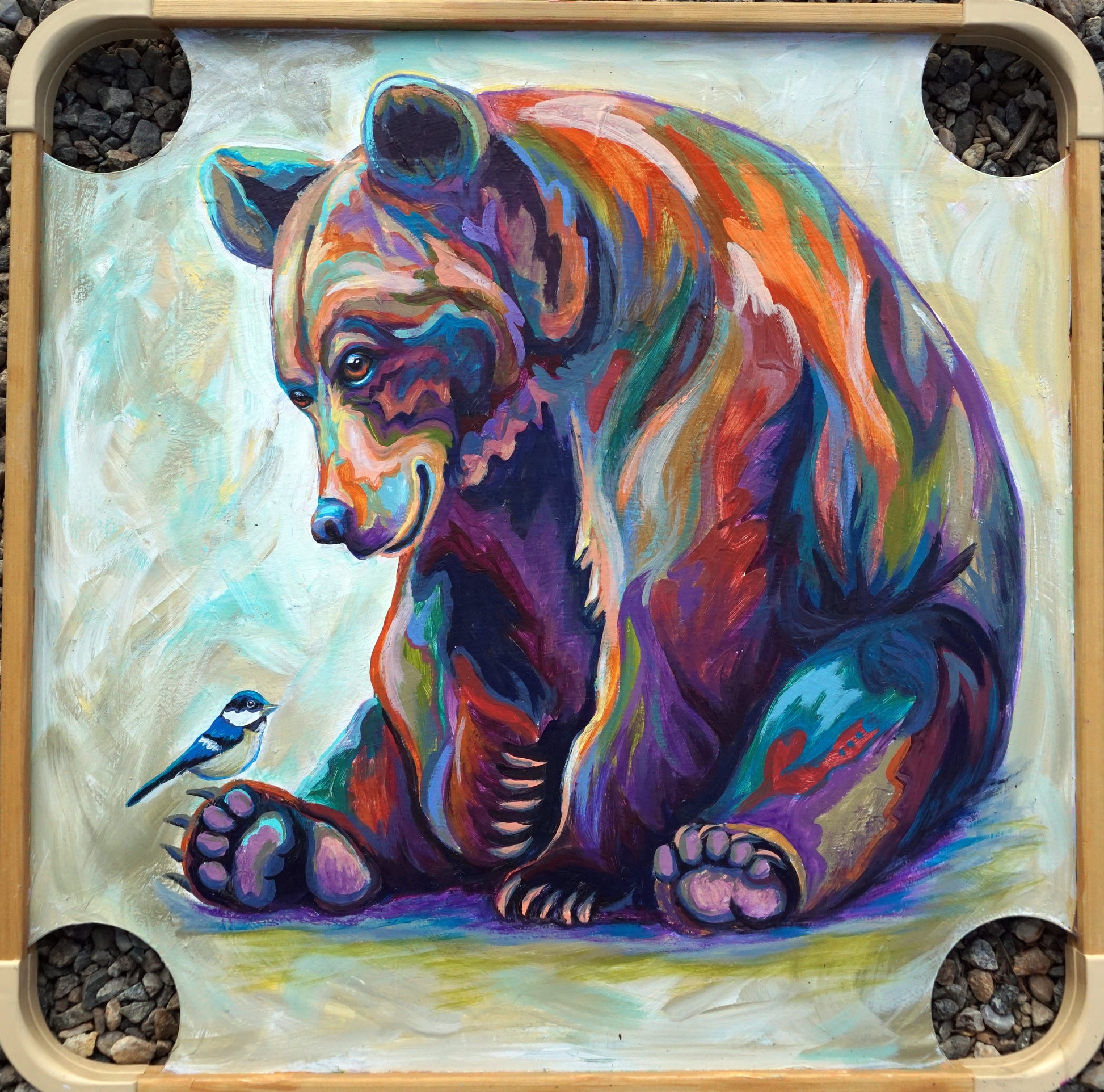 Original painting of a colorful grizzly bear leaning down toward a small mountain chickadee perched near his paw, painted on a reclaimed wooden carrom game board with corner pockets